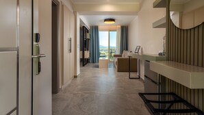 A modern hotel room with a large window, a sofa, a small table, and a mirror. - Corfelia Seaview Suites (Corfu)