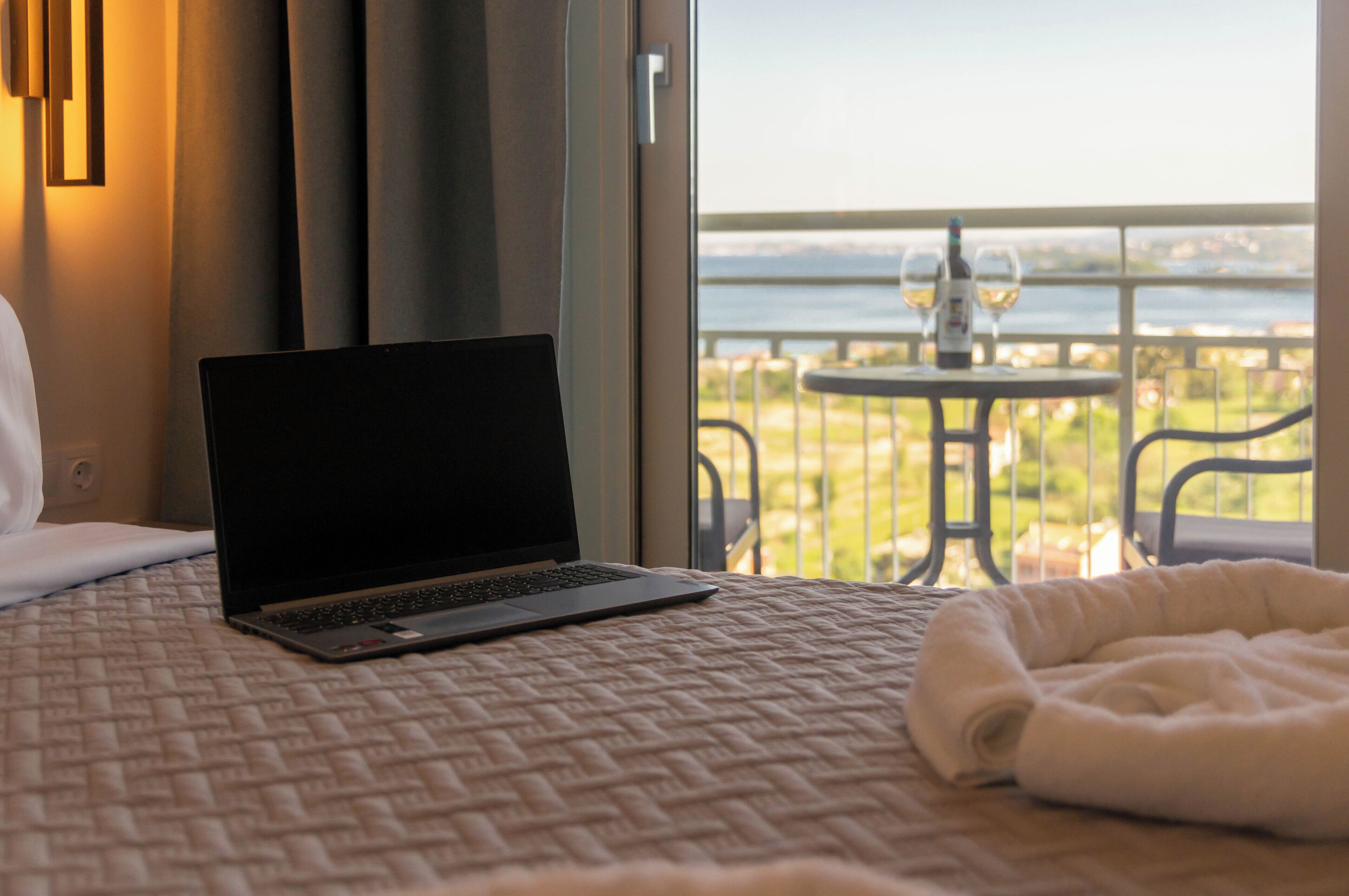 Una laptop sobre la cama, con vistas a un balcón y una mesa con vasos y una botella.