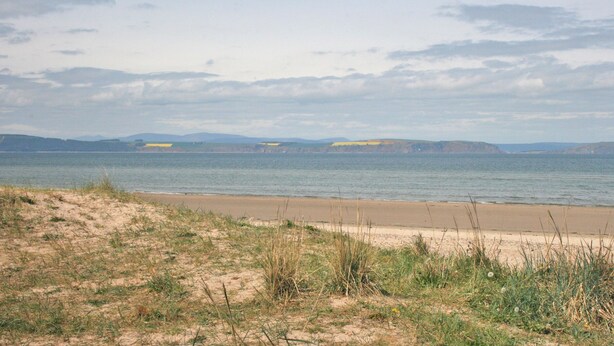 Un paysage côtier avec une plage de sable, des dunes herbeuses et une mer calme.