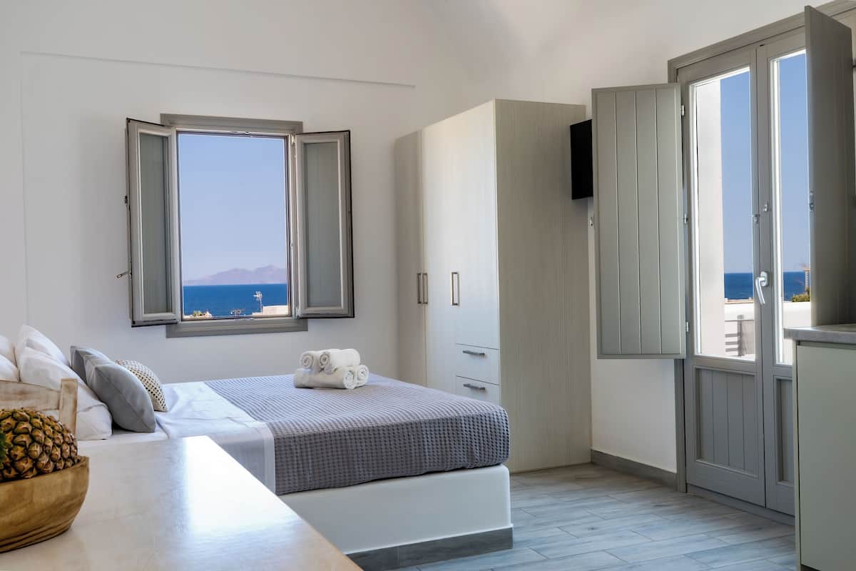 A bedroom with a bed, a wooden table with a bowl, a window with a view of the sea, and a wardrobe.