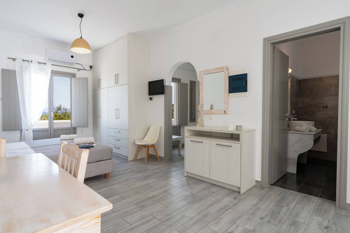 A modern, well-lit room with a dining area, a sofa, a TV, and a bathroom with a sink and mirror.
