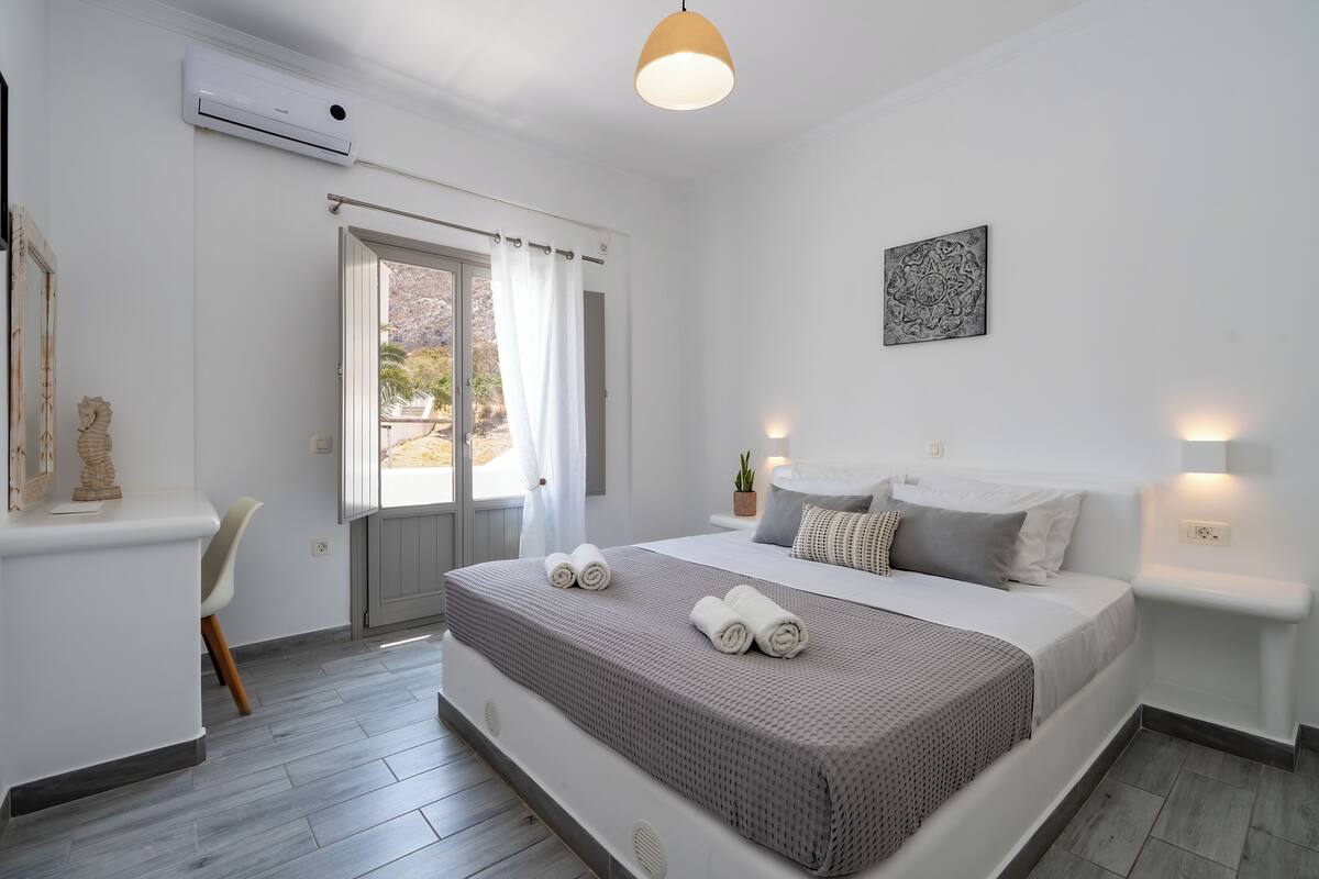 A modern bedroom with a bed, bedside tables, a window with curtains, an air conditioning unit, and a wall-mounted artwork.