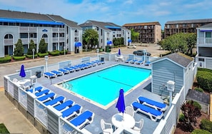 Una piscina con sillones y sombrillas, rodeada de edificios de varios pisos con balcones.