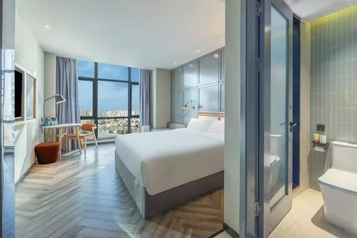 A modern hotel room with a large bed, a desk with a chair, a view of the city, and a separate bathroom area.