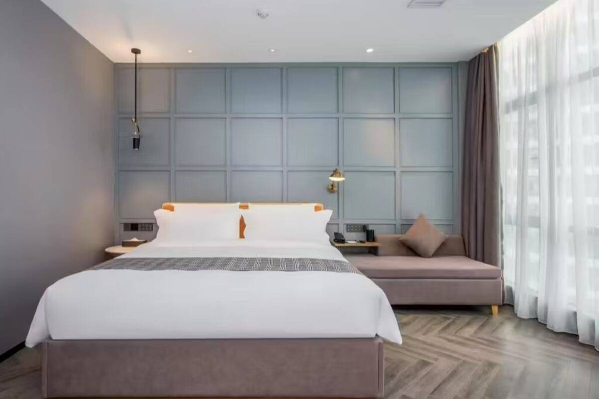 A modern hotel room with a large bed, a sofa, a desk, and a view of the city through large windows.