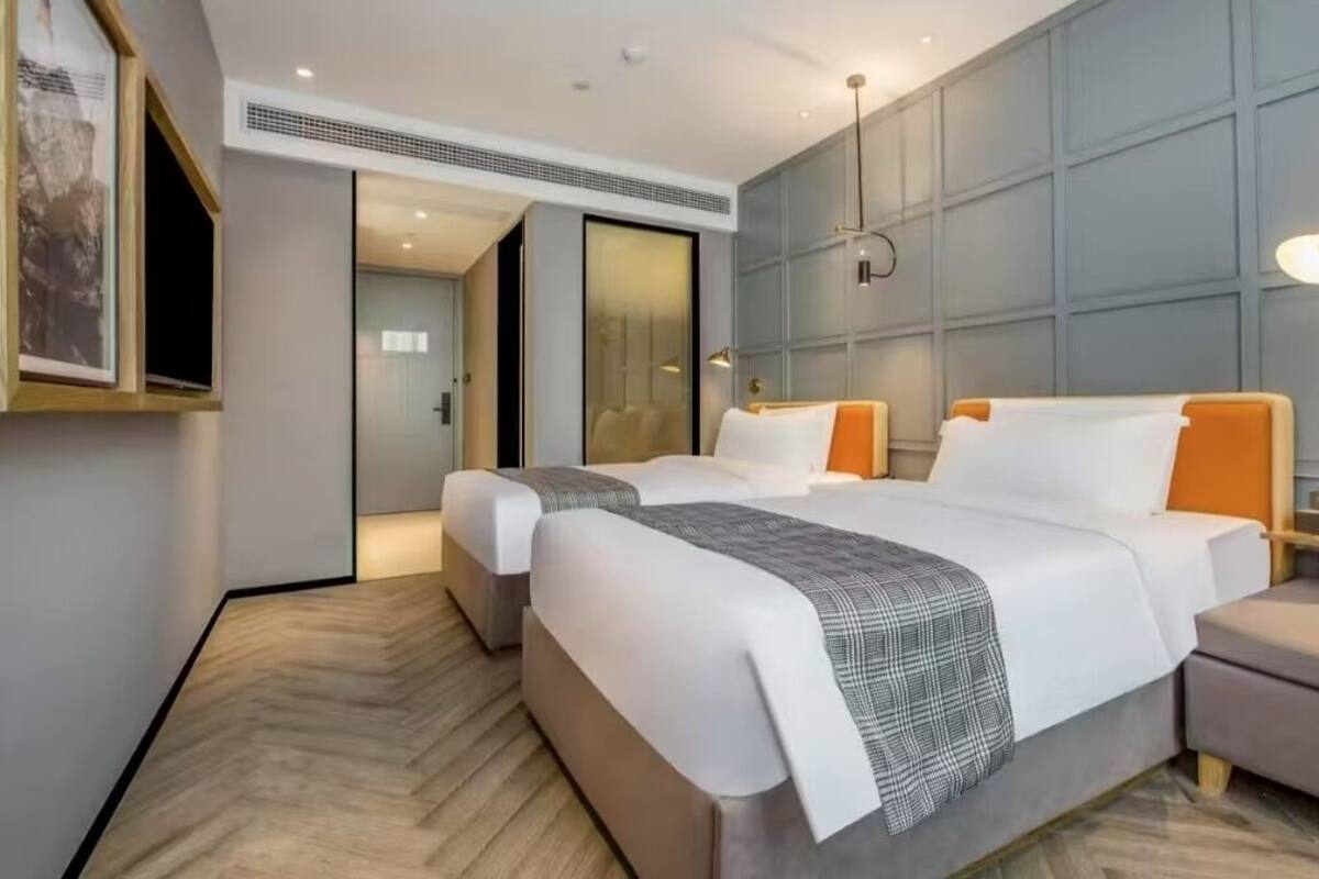 A modern hotel room with two beds, a large mirror, a nightstand, and a wall-mounted lamp.