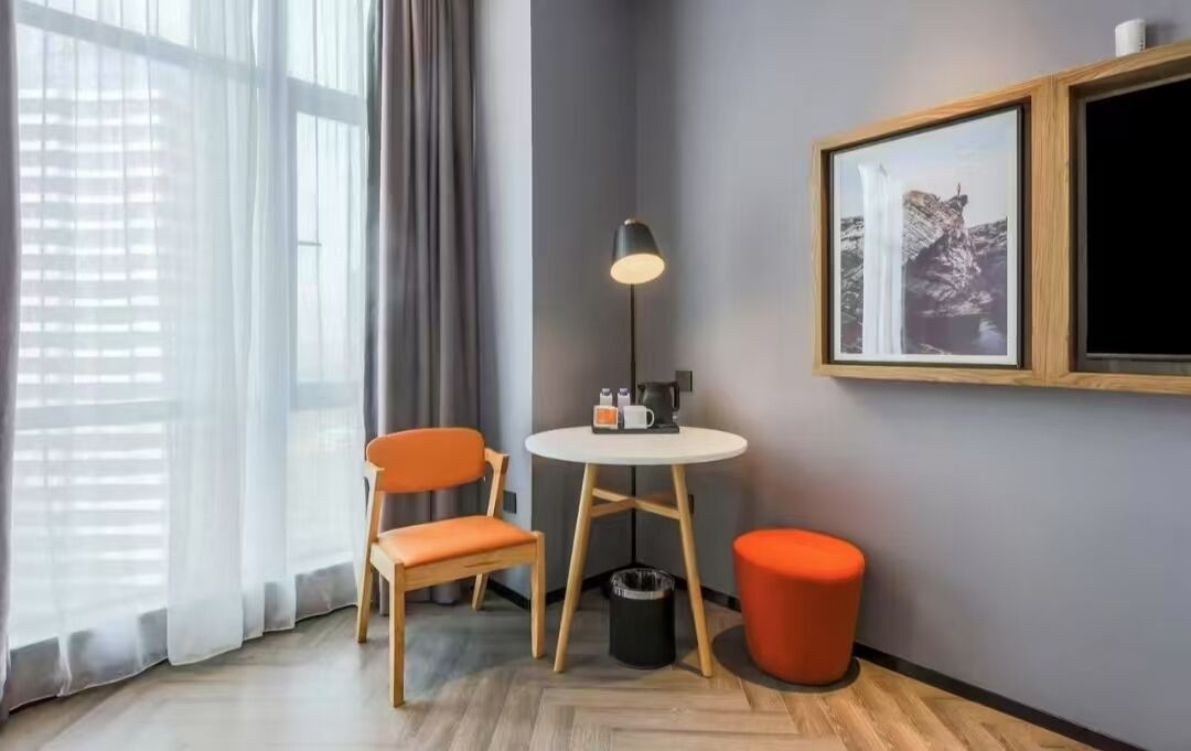 A corner room with a wooden floor, a round table with a lamp, two orange chairs, a small orange ottoman, and a framed picture on the wall.