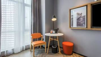 A corner room with a wooden floor, a round table with a lamp, two orange chairs, a small orange ottoman, and a framed picture on the wall.