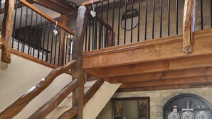 Una sala de estar acogedora con una escalera de madera, un sofá blanco con cojines estampados, un aparador de madera con platos decorativos y una planta en maceta sobre una mesa de madera.