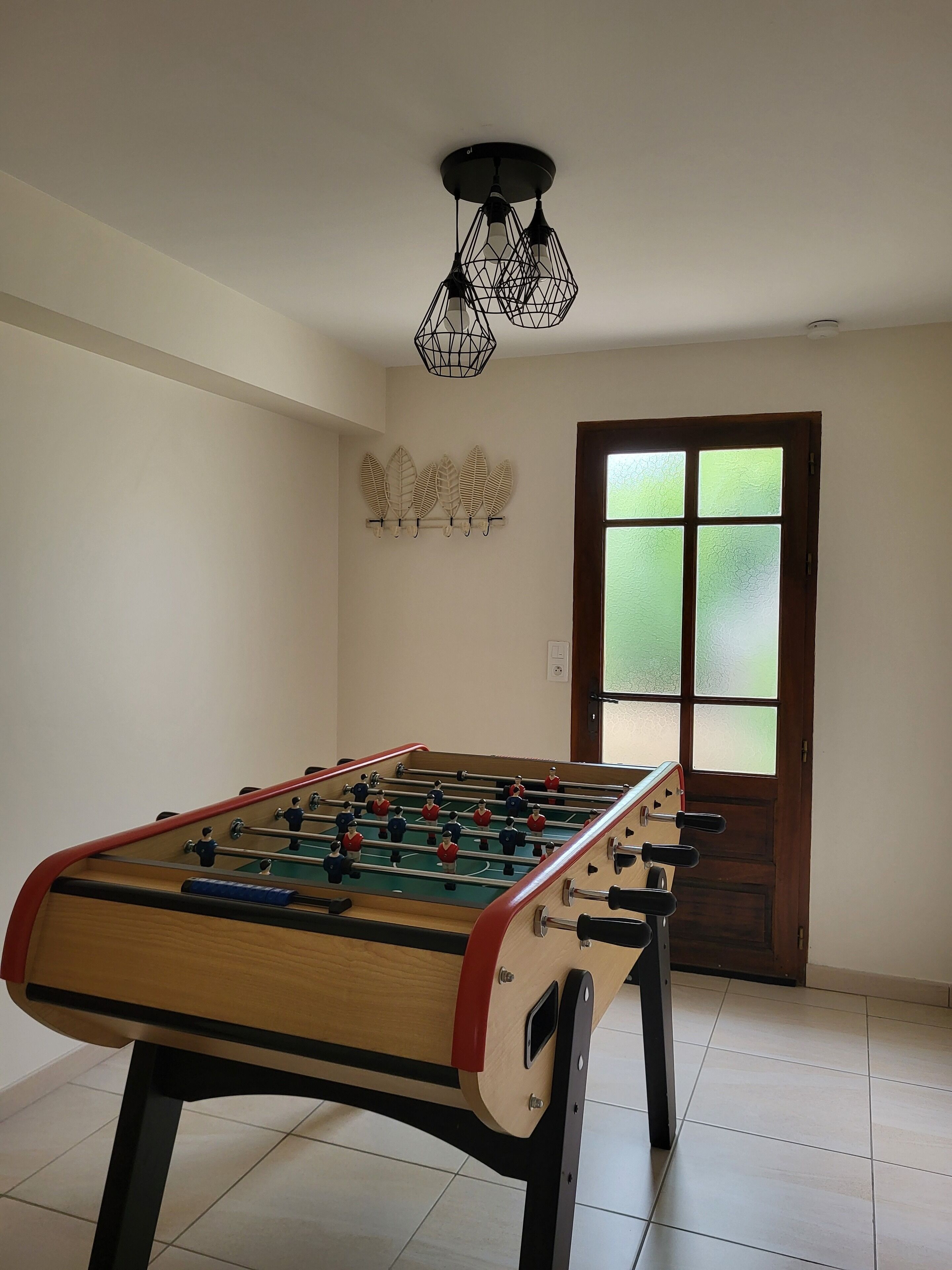 A foosball table in a room with a tiled floor, a wooden door with frosted glass panels, and a ceiling light fixture.