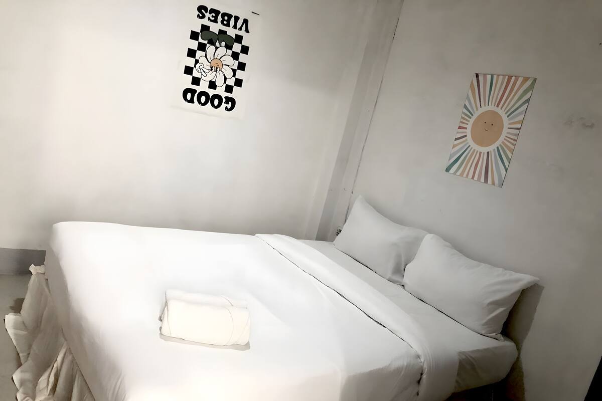 A single-bed room with white bedding, a wall-mounted poster, and a sun decoration.