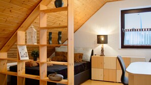 A loft bedroom with a sloped ceiling, wooden beams, a bed, a desk, and a window with curtains.