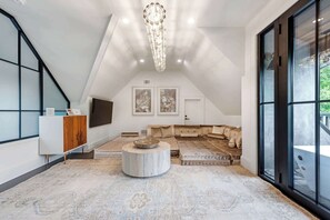 A spacious loft with a high ceiling, a large window, a modern chandelier, a sofa set, and a television.