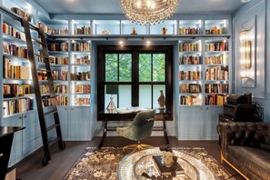 Ein Zimmer mit einem großen Bücherregal voller Bücher, einem Schreibtisch mit Lampe, einem Sessel, einem Ledersofa und einer Kandelaber.