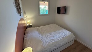 Un dormitorio con cama, lámpara de noche, televisión montada en la pared y ventana con vista a follaje.