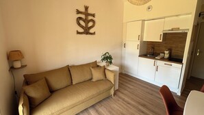 Una sala de estar con un sofá, una mesita de noche con una lámpara, una planta en maceta y una cocina con armarios y un lavamanos.