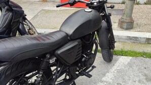 Une moto est garée sur une surface pavée, avec un fond rouge et une clôture métallique en arrière-plan.