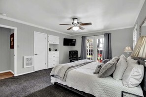 Un dormitorio con cama, ventilador de techo, televisión, ventana con cortinas y una puerta.