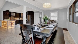 Un espace repas avec une longue table, des chaises, un lustre et une cuisine en arrière-plan.