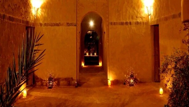 Un couloir faiblement éclairé, avec une arche centrale menant à une pièce, le tout bordé de murs aux motifs texturés et de petites lumières disposées le long du passage.