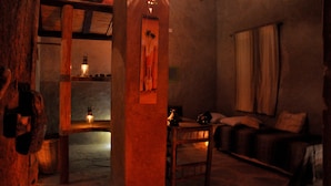 Une pièce faiblement éclairée, avec un plafond en bois, un canapé, une table et une étagère fixée au mur, sur laquelle se trouvent des livres et une petite lampe.