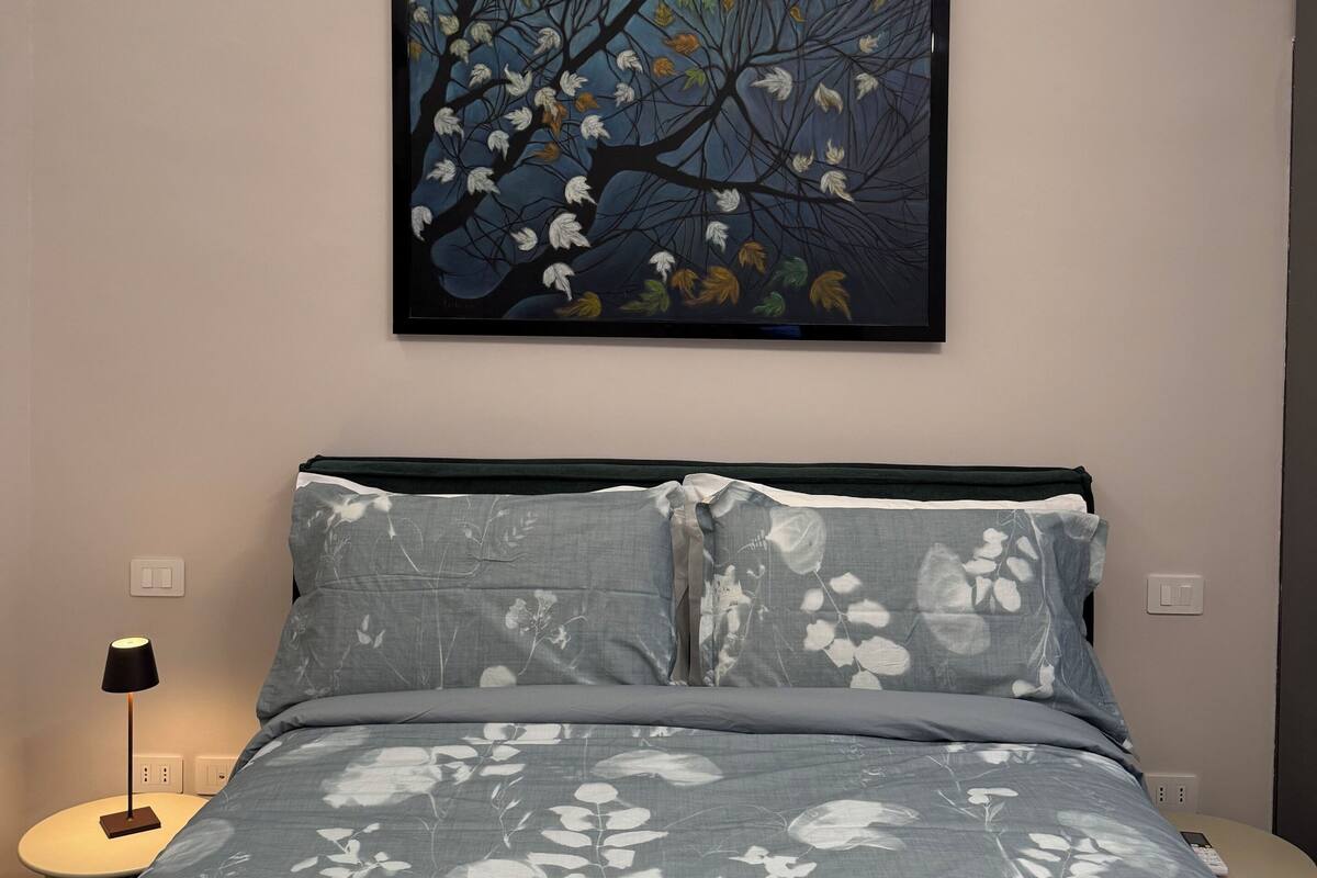 Un dormitorio con cama, mesita de noche, lámpara y un cuadro colgado en la pared.