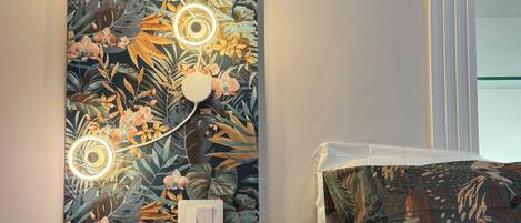 A room with a floral wallpaper, a bed with a patterned pillow, a bedside table, and a wall-mounted light fixture.