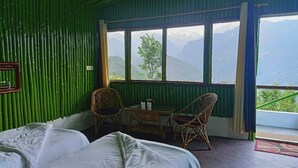A room with a bed, two wicker chairs, a small table, and a view of mountains through large windows.