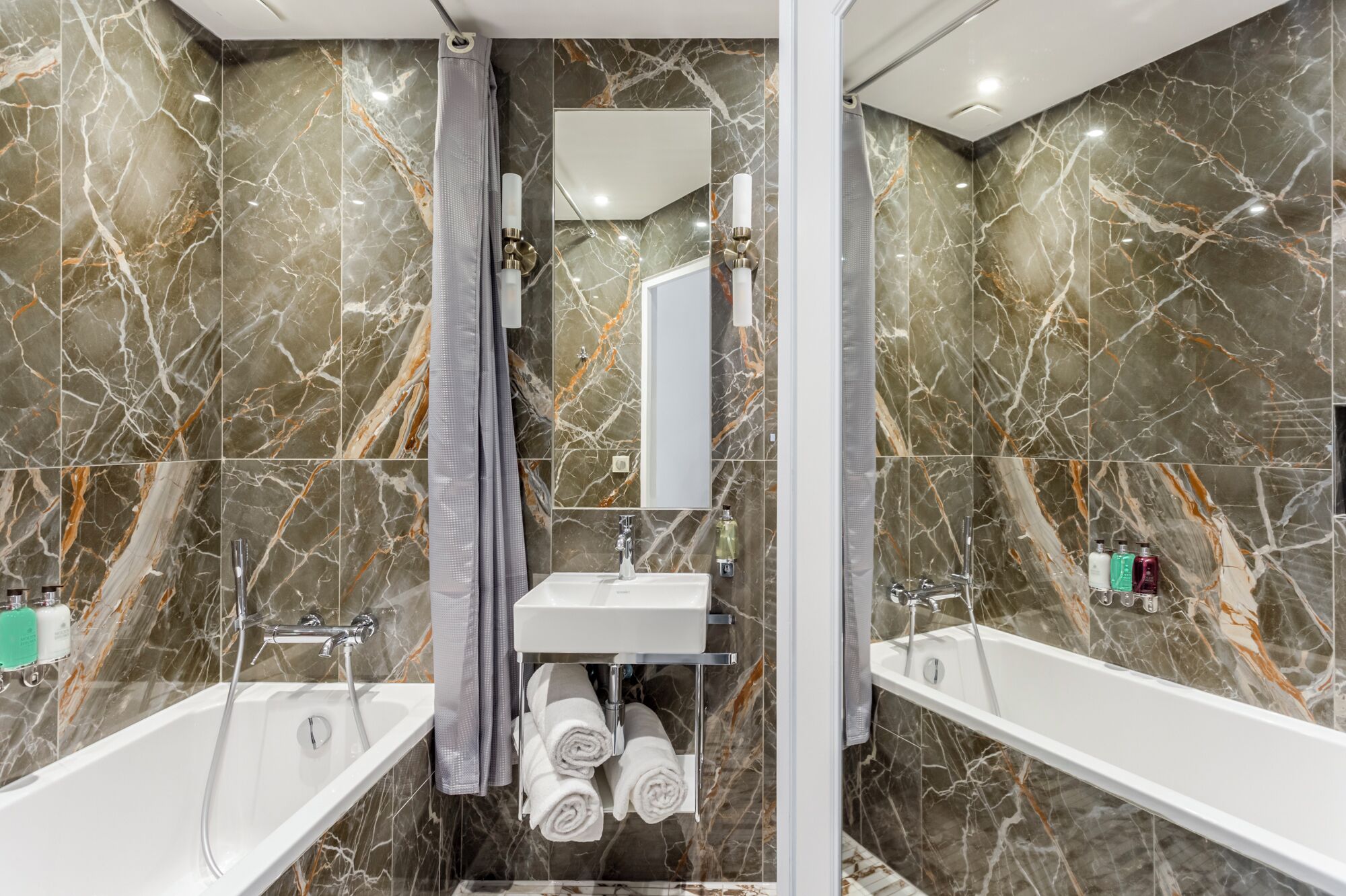 A modern bathroom with marble walls, a central sink, two side bathtubs, and a shower curtain.