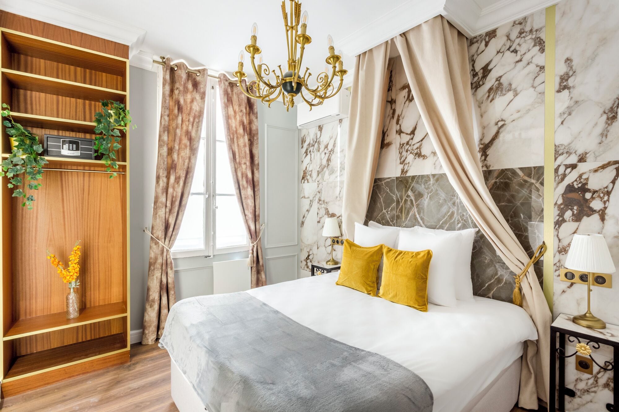 A bedroom with a bed, a bedside table, a nightstand, a chandelier, curtains, and a wooden wardrobe.