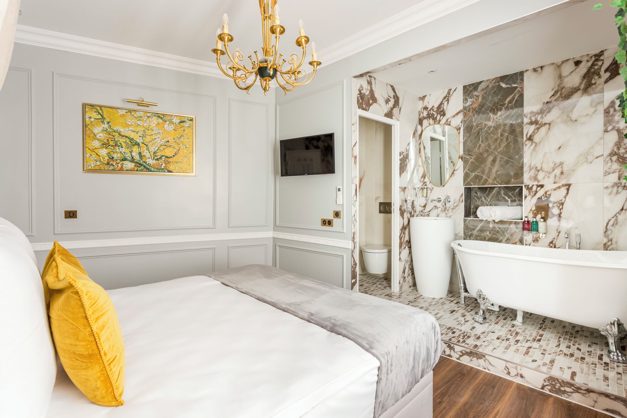 A hotel room with a bed, a large painting, a television, a marble bathroom with a bathtub, and a chandelier.
