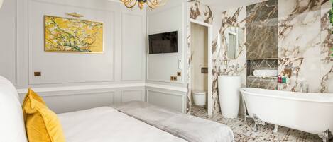 A hotel room with a bed, a large painting, a television, a marble bathroom with a bathtub, and a chandelier.