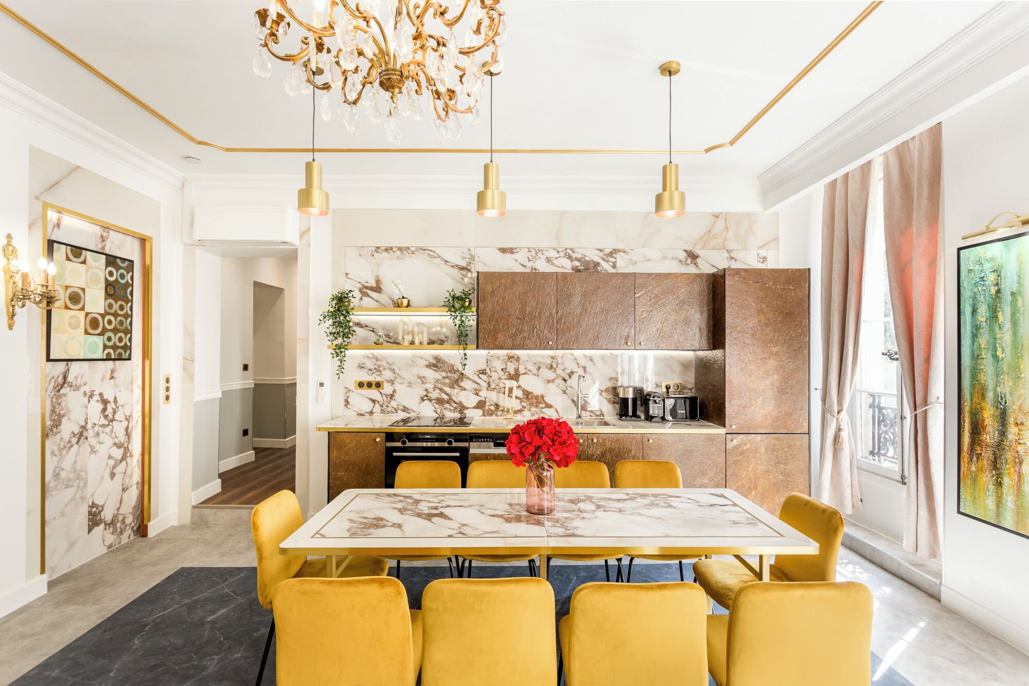 A dining area with a marble table, yellow chairs, a chandelier, and a kitchen with marble backsplash and wooden cabinets.