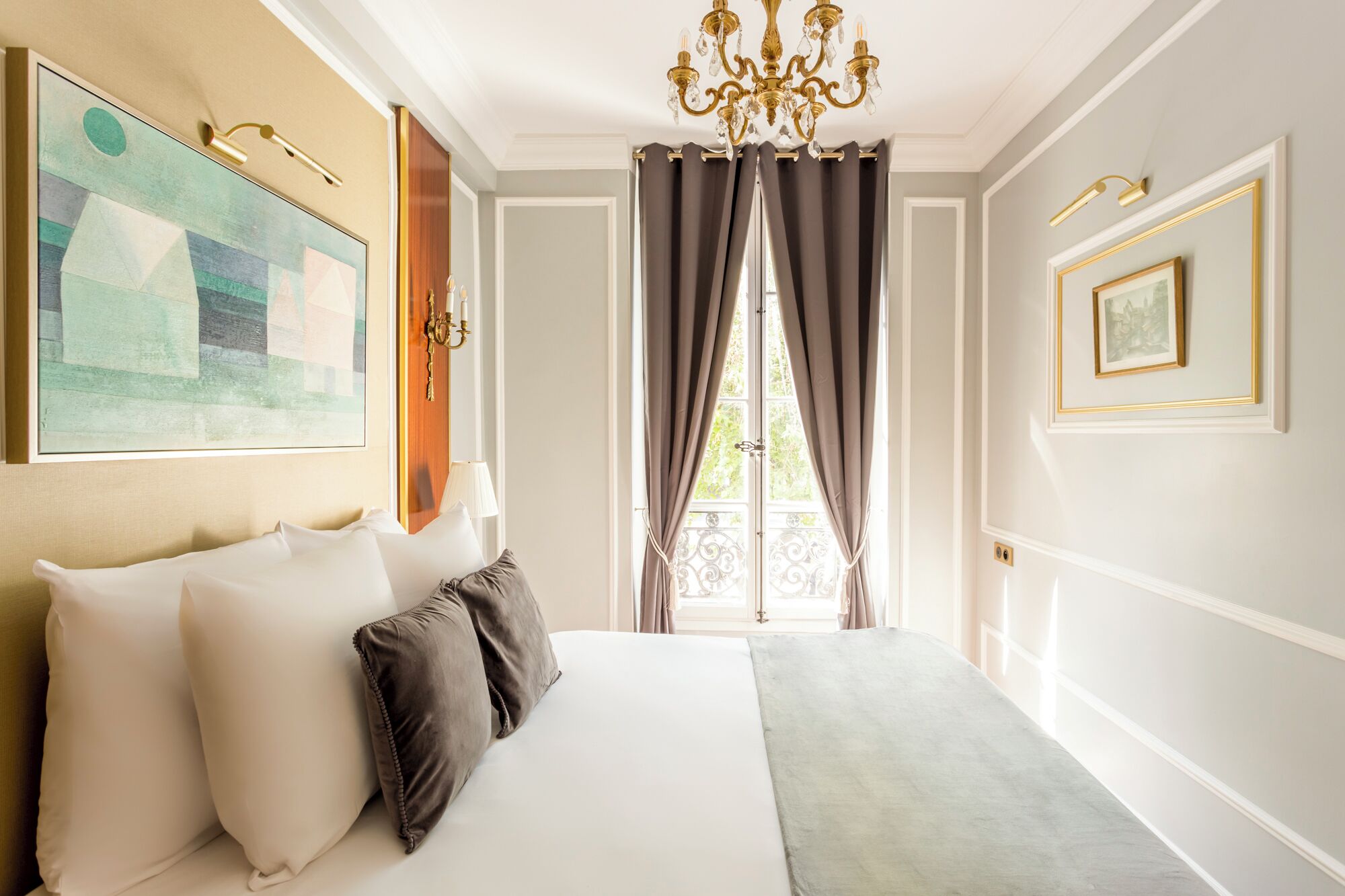 A hotel room with a large bed, a chandelier, a painting, and a window with curtains.