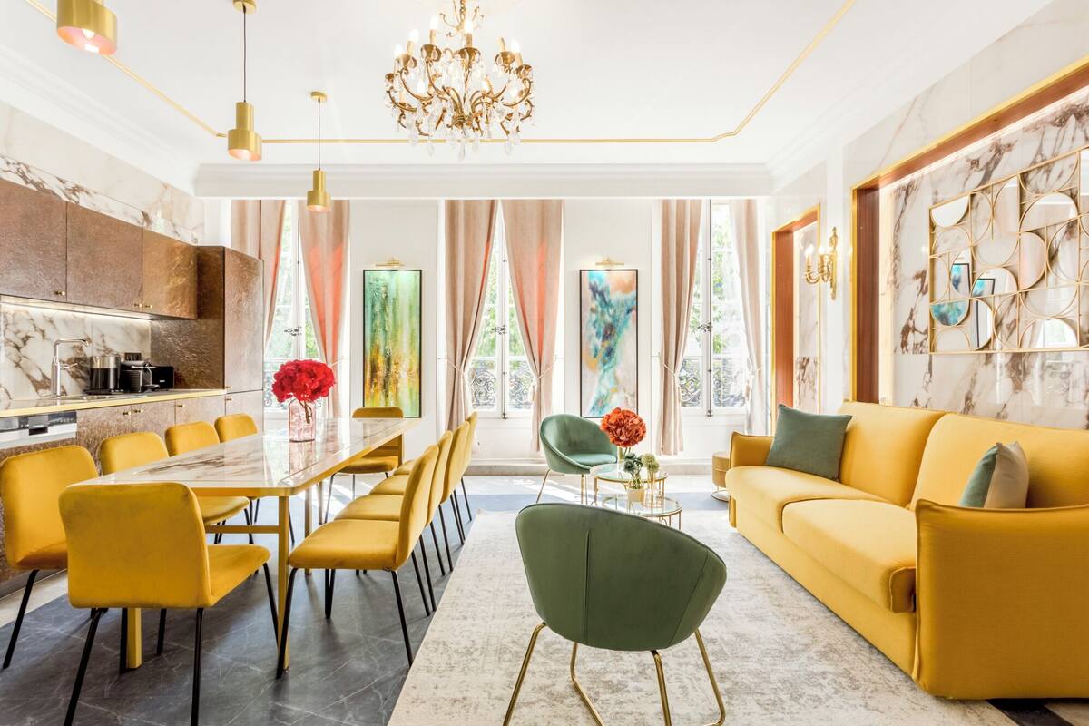 A modern living room with a yellow sofa, green armchair, and a marble dining table with yellow chairs.