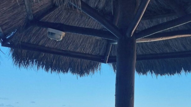 Un patio frente a la playa con un toldo de paja, dos sillas y vista al océano y un barco a lo lejos.