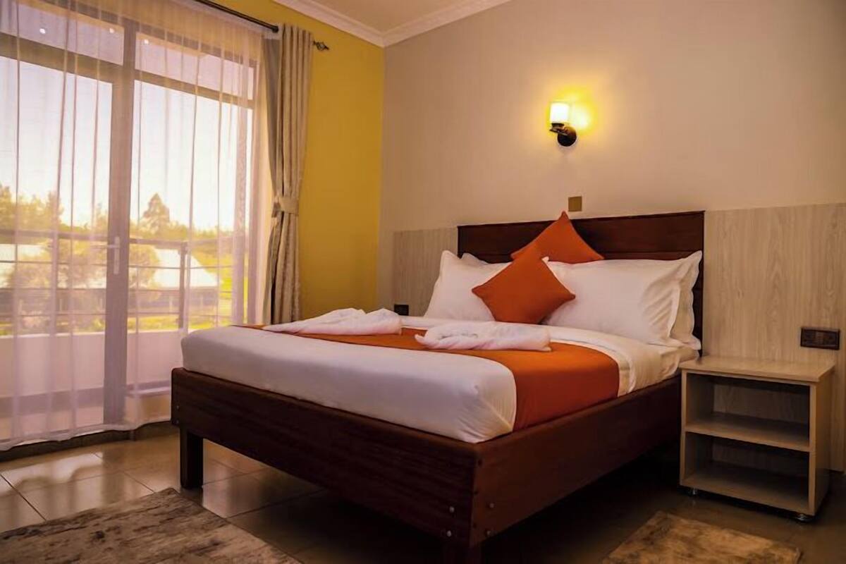 A hotel room with a bed, a nightstand, a wall-mounted light, and a view through a window with curtains.