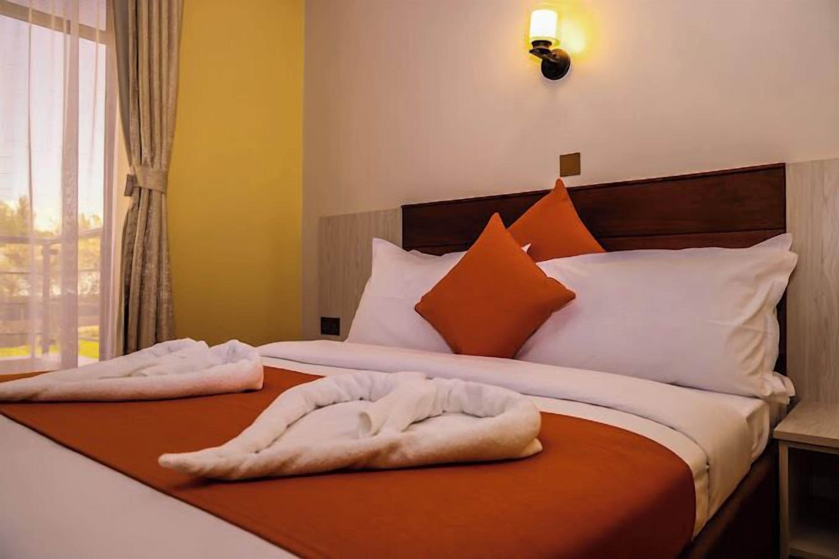 Habitación de hotel con cama, almohadas anaranjadas, ropa de cama blanca, una mesita de noche y una lámpara de pared.