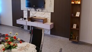 A modern living room with a flat-screen TV mounted on a marble wall, a wooden shelf with decorative items, and a dining area with a table set for two. - Furnished studio, Douala-Total Nkolbong close to the road and all amenities (Douala)