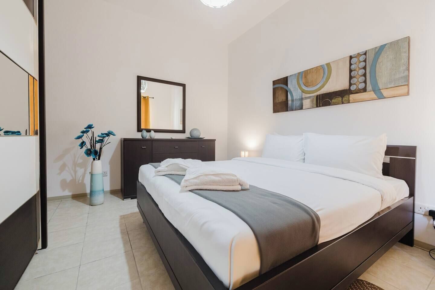 A modern hotel room with a large bed, a dresser, a mirror, and a painting on the wall.