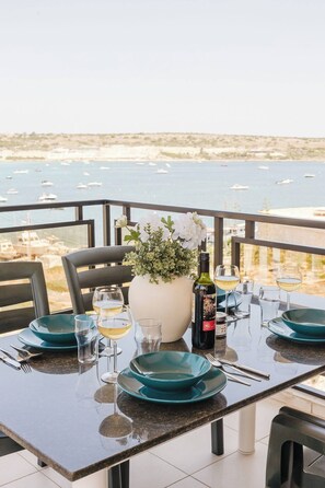 A dining table set with teal plates, wine glasses, and a bottle of wine, overlooking a marina with boats. - Spacious seafront apartment in Għadira Bay with 3 bedrooms, 2 bathrooms, and a large terrace boasting spectacular sea views. Located on the third floor (no lift) and only 1 minute from the sandy beach — perfect for up to 5 guests. (Il-Mellieħa)