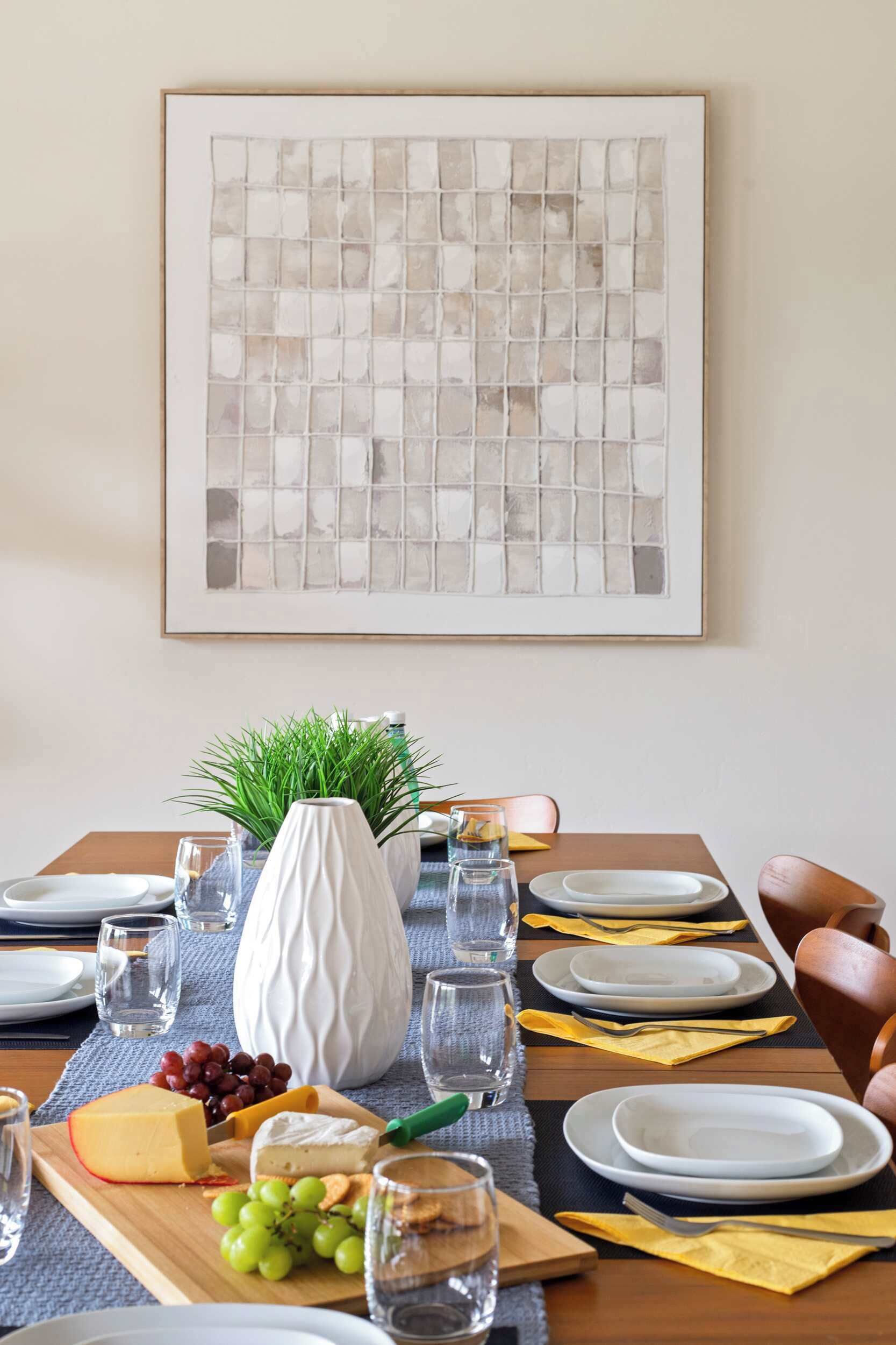 Una mesa de comedor dispuesta con platos, vasos, un florero y una tabla de quesos.