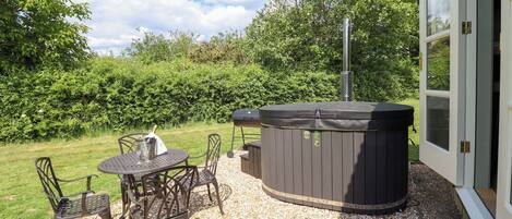 A hot tub with a chimney, surrounded by a patio with chairs and a table, set against a backdrop of trees and a house.
