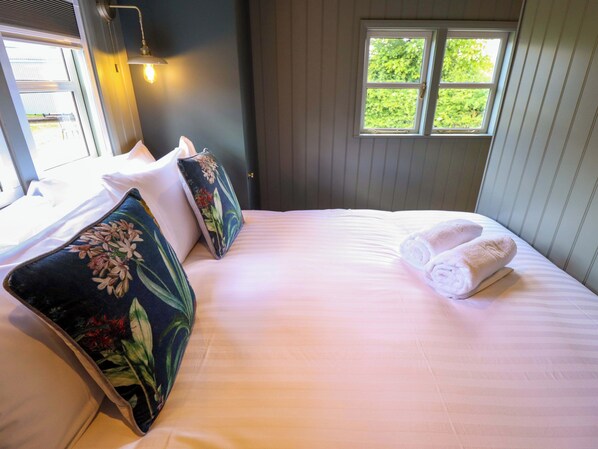 A neatly made bed with floral pillows and a rolled towel, a window with a view of greenery, and a wall-mounted light fixture. - Bluebell (Grimsby)