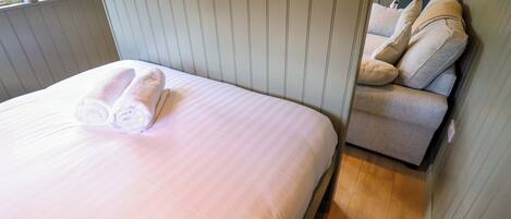 A neatly made bed with rolled white towels, a wooden floor, and a built-in sofa with cushions.