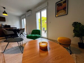 Una sala moderna con una mesa de madera, un sillón verde y una silla amarilla. Hay un ventanal con vista a zonas verdes.