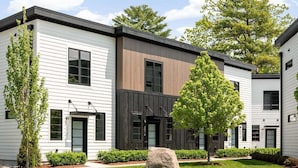 A modern multi-story building with a combination of white and dark wood paneling, large windows, and a well-maintained lawn with trees and a rock feature. - Hamilton Place - 305 (3 units) Sleeps 12 (Traverse City)