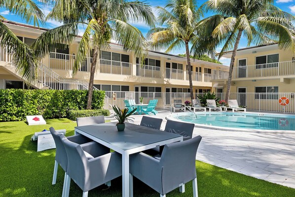 Zona de piscina con mesa y sillas, una piscina y un edificio con balcones y palmeras.