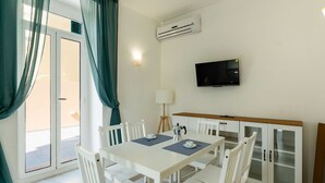 Un comedor con una mesa y sillas blancas, un televisor montado en la pared y un equipo de aire acondicionado.