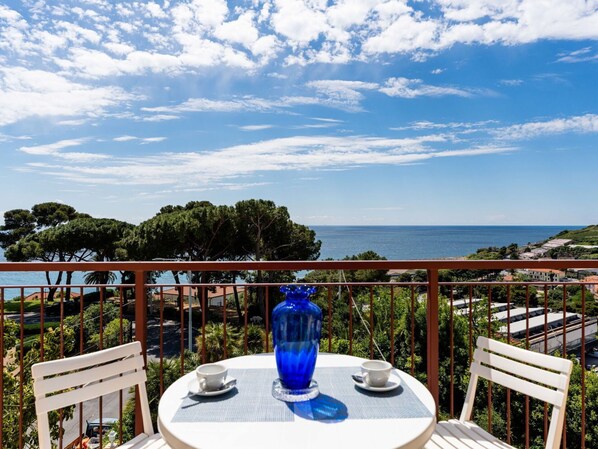 Una mesa redonda con dos sillas blancas, un jarrón azul y dos tazas blancas en un balcón con vistas a la costa, árboles y edificios.